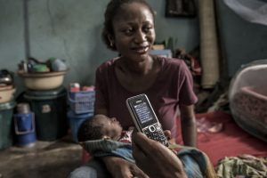 Woman sitting holding baby, with someone holding a cellphone